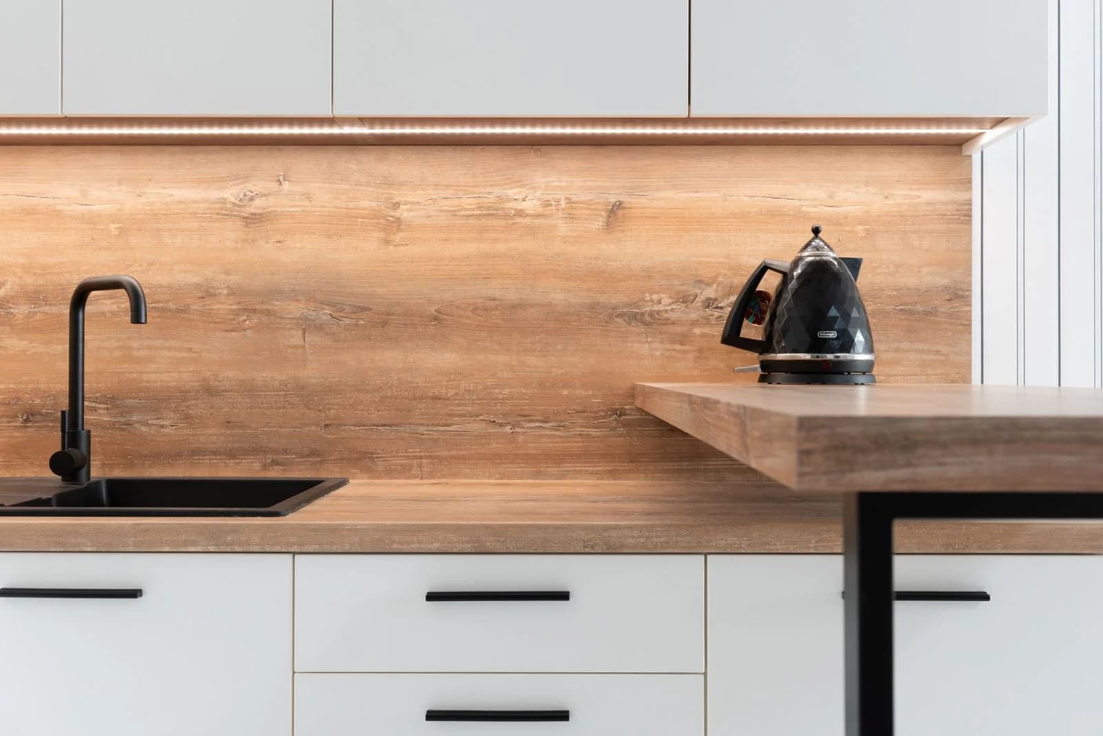 Detalle de cocina con mesada y pared de madera, muebles blancos y grifería negra.